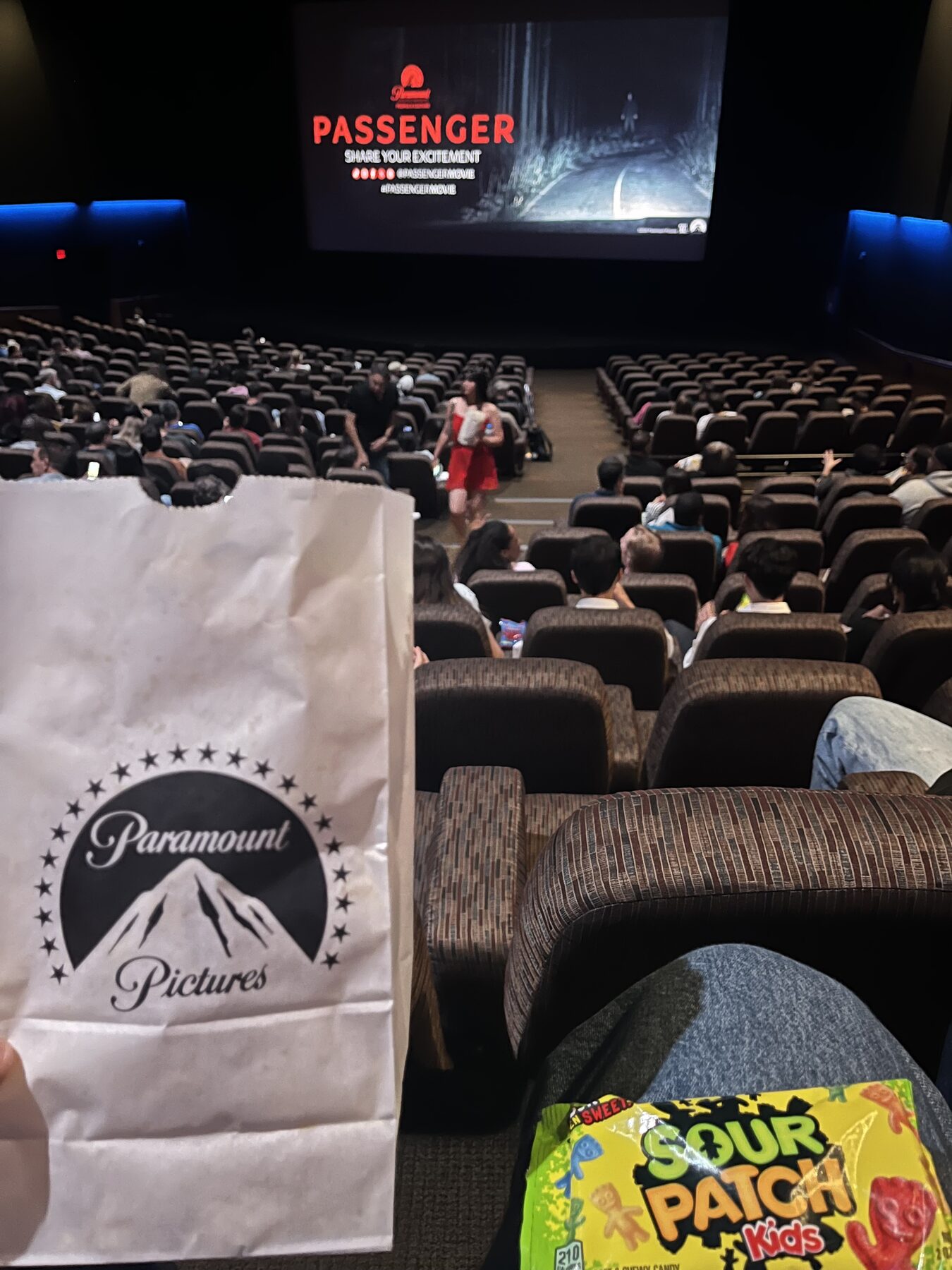 Inside the screening — Paramount Pictures popcorn bag held up, Passenger title card on screen, Sour Patch Kids on lap