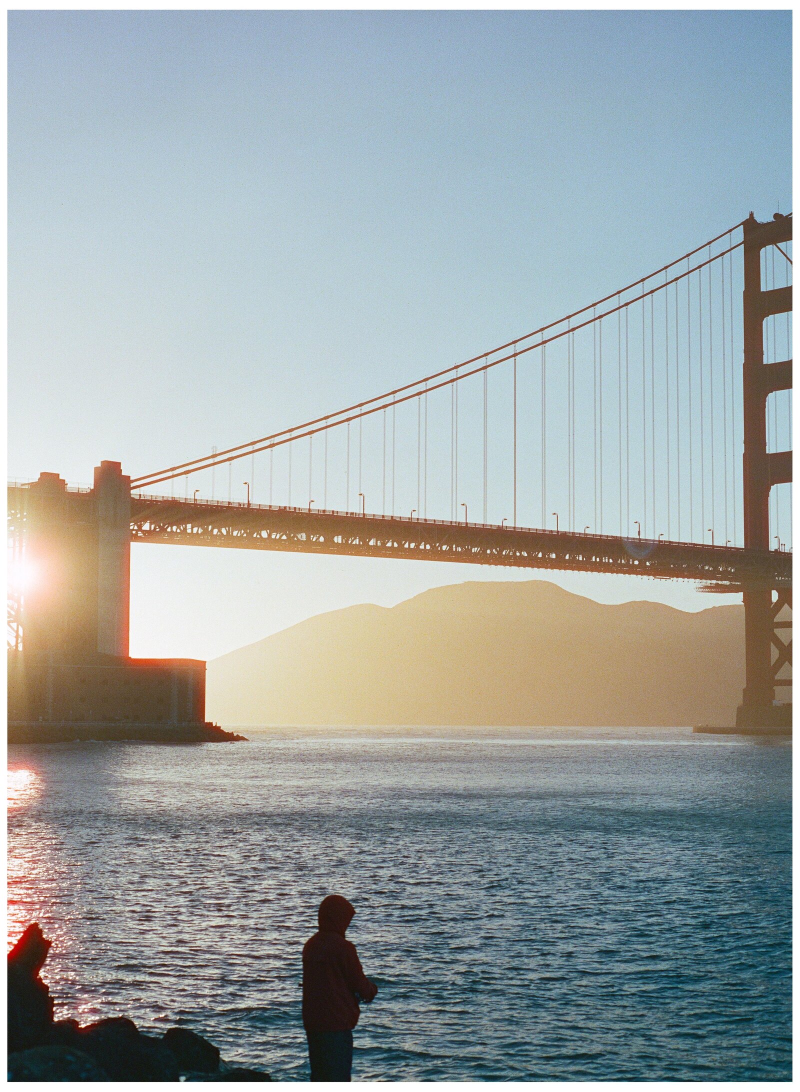 The Golden Gate Bridge at golden hour on film — the sun flaring behind the south tower, and a hooded figure standing alone at the water's edge below the span